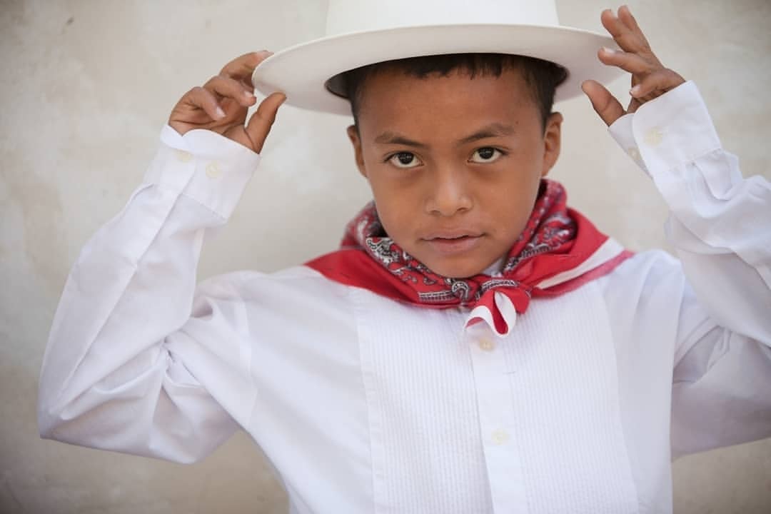 Portrait of a young boy wearing a white shirt, red neckerchief, and white hat, looking directly at the camera with a neutral expression.