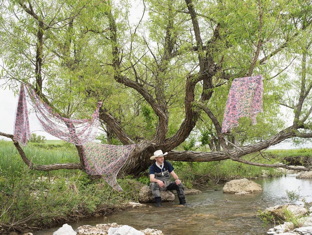 Man sits in a stream beneath a willow tree draped with floral fabric; rural scene with grassy banks and overcast sky.