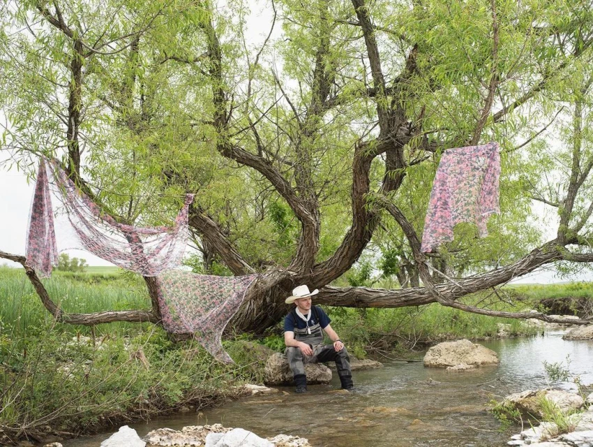 Man sits in a stream beneath a willow tree draped with floral fabric; rural scene with grassy banks and overcast sky.