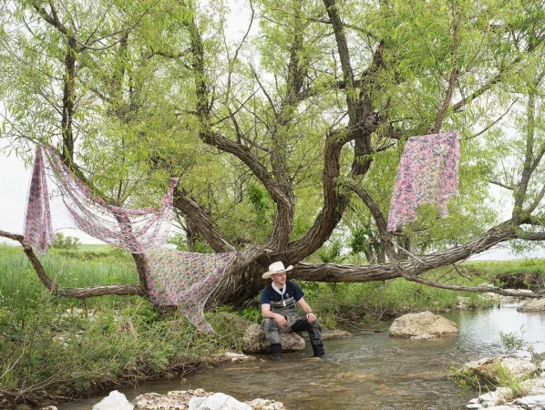 Man sits in a stream beneath a willow tree draped with floral fabric; rural scene with grassy banks and overcast sky.