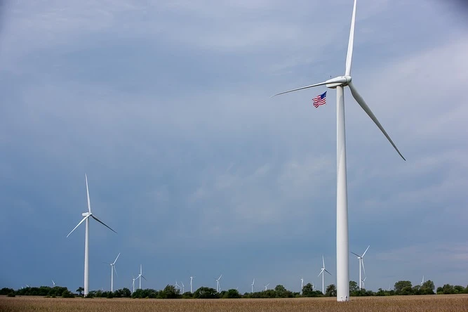 Wind turbines stand in a field with the American flag attached to one against a cloudy sky, showcasing renewable energy.