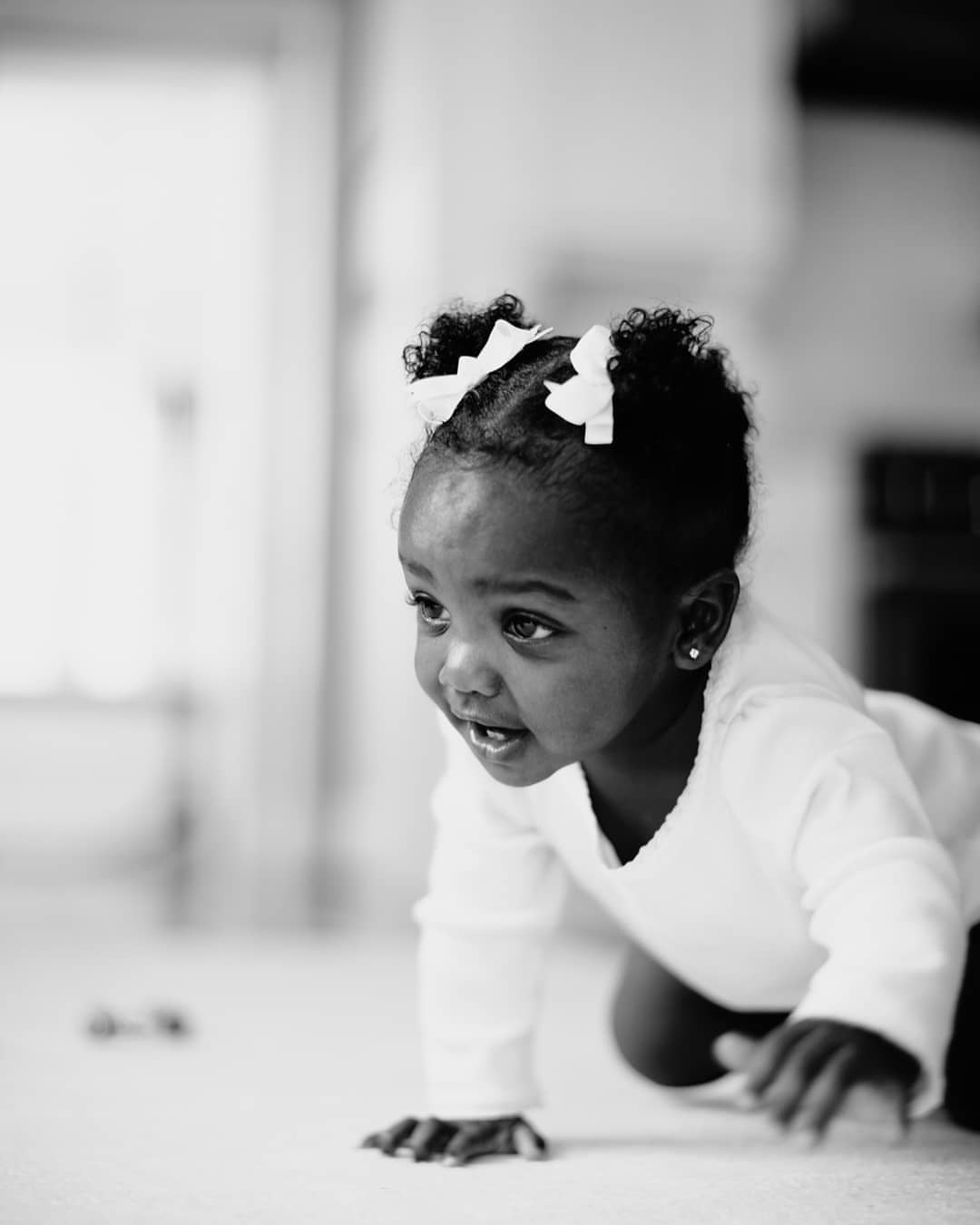 Black and white portrait of a baby crawling on the floor indoors, wearing a white shirt and matching hair bows, in a shallow focus.
