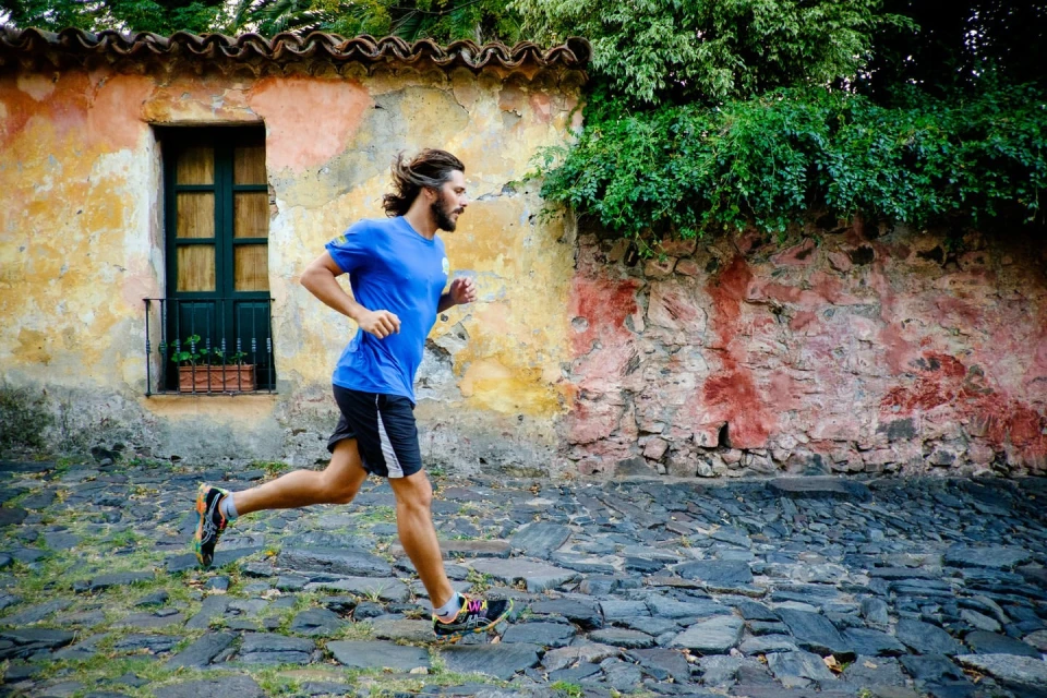 Man in blue shirt and black shorts runs on cobblestone street next to aged, colorful building under tree shade; athletic activity.