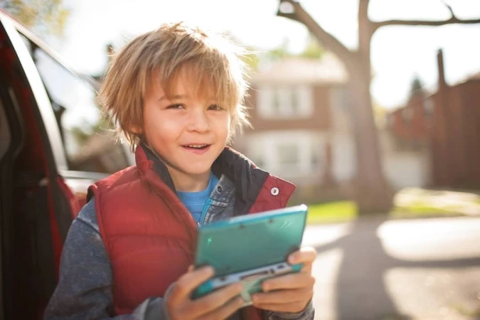 Young boy in red vest holds a blue portable game console near a red car and brick buildings on a sunny day outdoors.