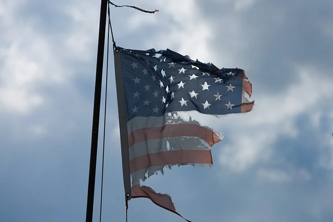 Tattered United States flag waving against cloudy sky. Weathered American flag with stars and stripes on a metal pole outdoors.