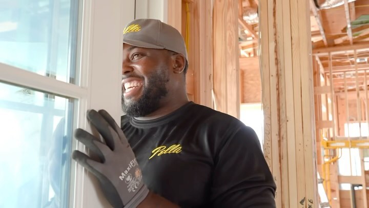 Adult Black man wearing Pella shirt and gloves installs window in home construction, smiling with a happy expression in sunlight.