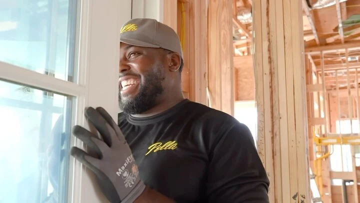 Adult Black man wearing Pella shirt and gloves installs window in home construction, smiling with a happy expression in sunlight.