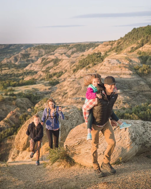 A family hikes in a desert environment, with a father carrying a child piggyback, another child hiking with trekking poles, and a woman hiking in the background.