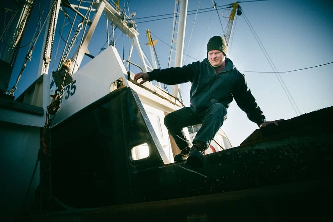 Adult male wearing a beanie, balancing and jumping from one fishing boat to another on a sunny day; close-up, eye-level angle.
