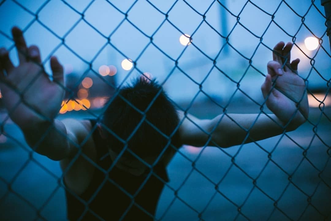 Person with dark hair and a black shirt grips a wire fence in a dark urban setting, city lights blurred in the background.