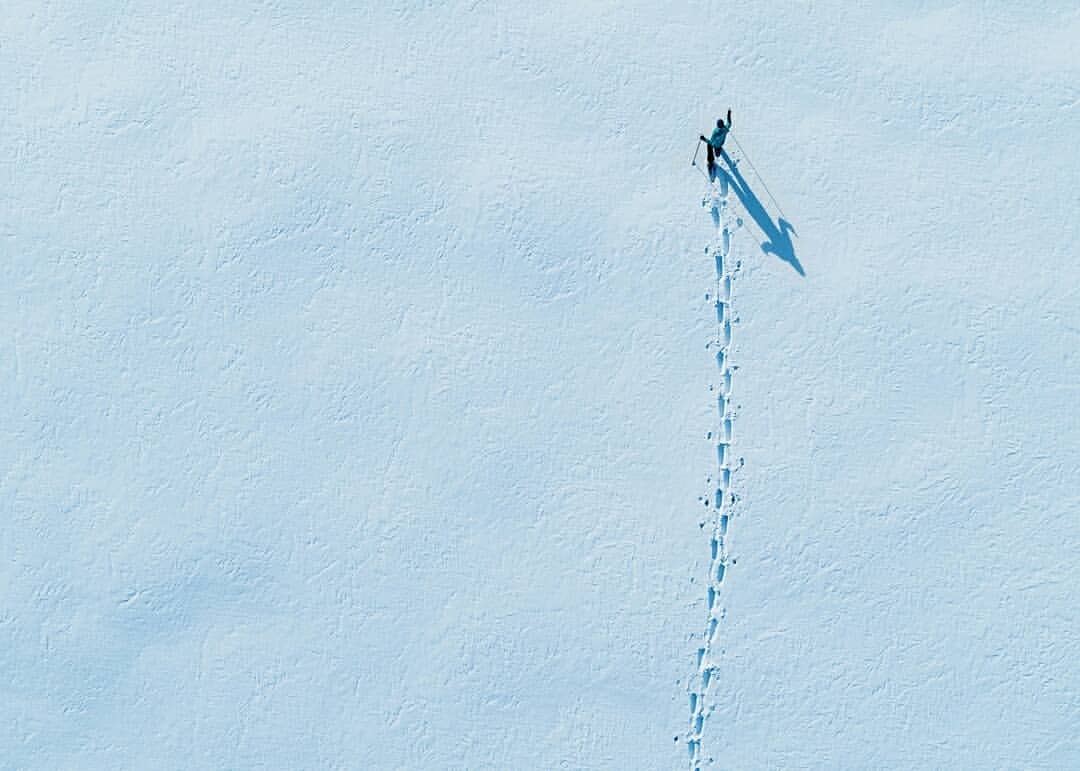 Aerial view of a lone person cross-country skiing in a snowy landscape, leaving a trail of footprints in the snow with a long shadow.