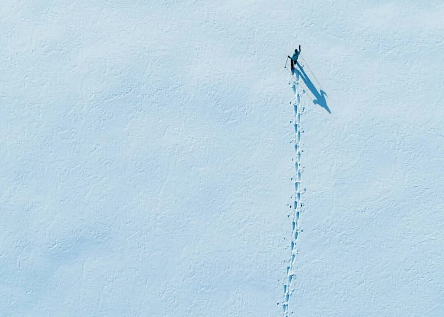 Aerial view of a lone person cross-country skiing in a snowy landscape, leaving a trail of footprints in the snow with a long shadow.