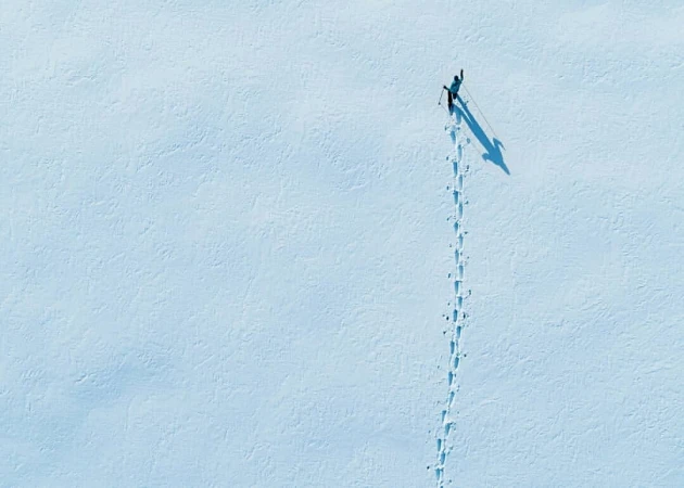 Aerial view of a lone person cross-country skiing in a snowy landscape, leaving a trail of footprints in the snow with a long shadow.