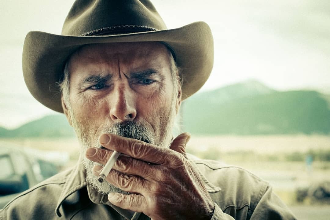 Close-up portrait of a weathered senior man in a cowboy hat smoking a cigarette outdoors with mountains in the background.