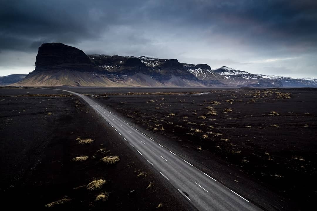 Road stretches through black sand landscape towards jagged mountain range under dramatic sky, Iceland. Moody scenic vista.