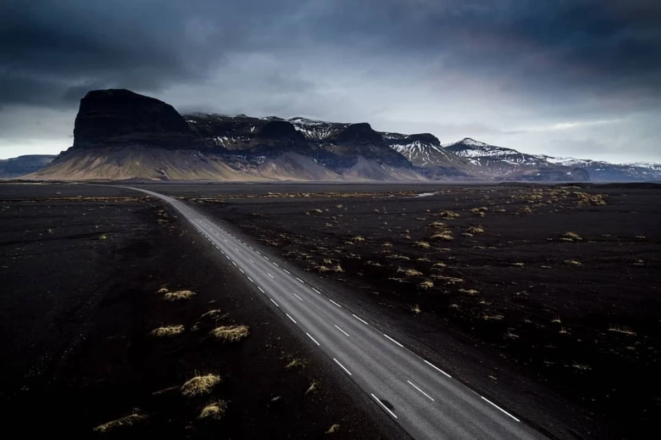 Road stretches through black sand landscape towards jagged mountain range under dramatic sky, Iceland. Moody scenic vista.