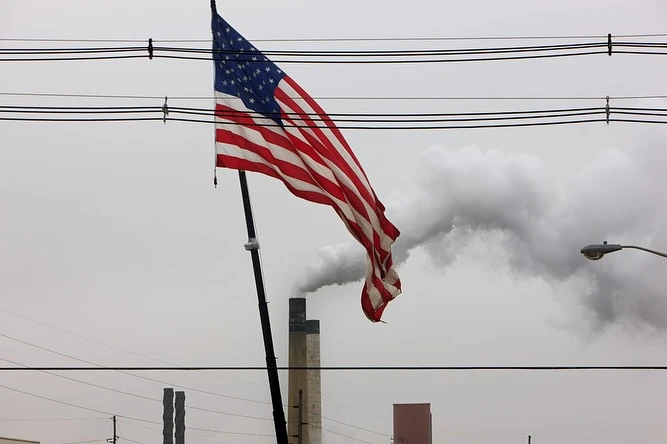 American flag waves in the wind against a backdrop of industrial smokestacks emitting smoke into a cloudy sky, near power lines.