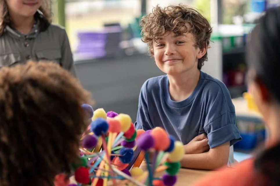 A boy with curly hair smiles in a classroom setting with colorful crafts, surrounded by other children. Educational environment.