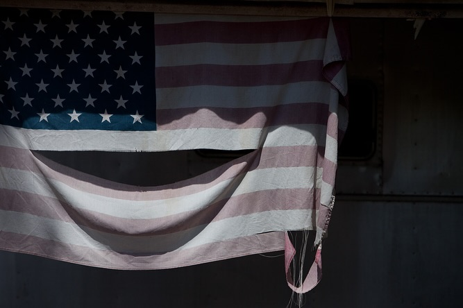 Faded and tattered United States flag hangs indoors with dramatic lighting, creating an evocative and somber display of patriotism.
