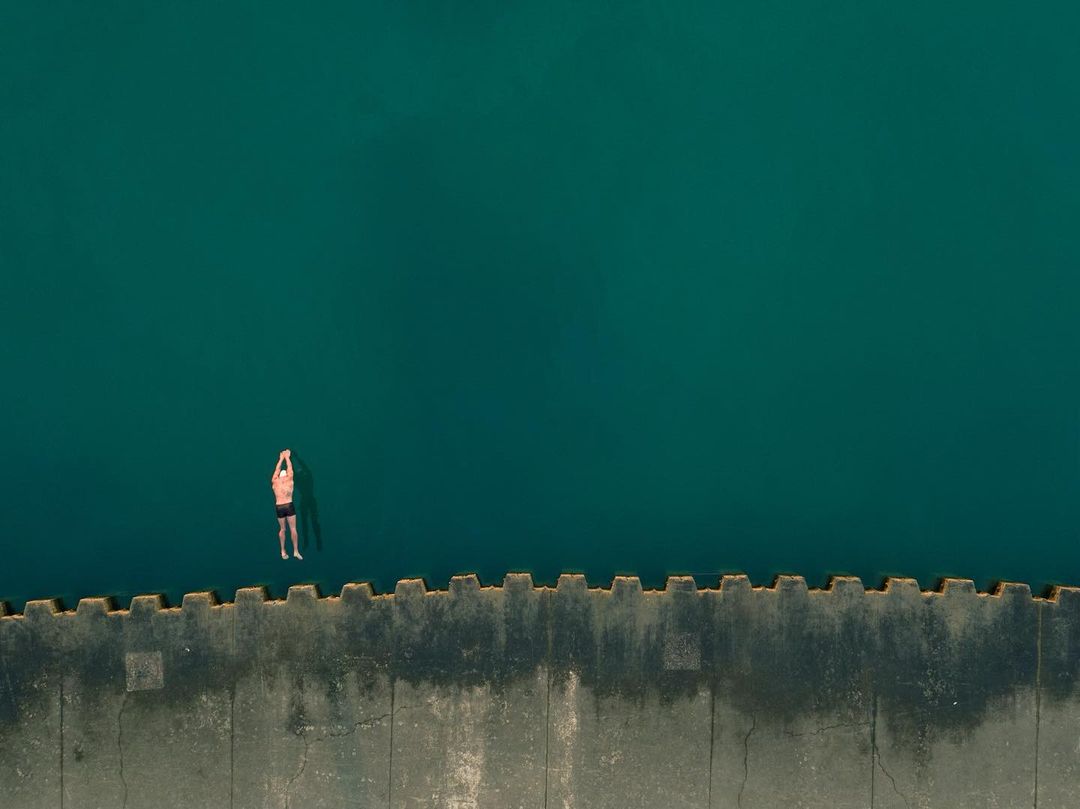 Aerial view of an adult male floating in turquoise water next to a concrete pier on a sunny day, showing tranquility and open water.