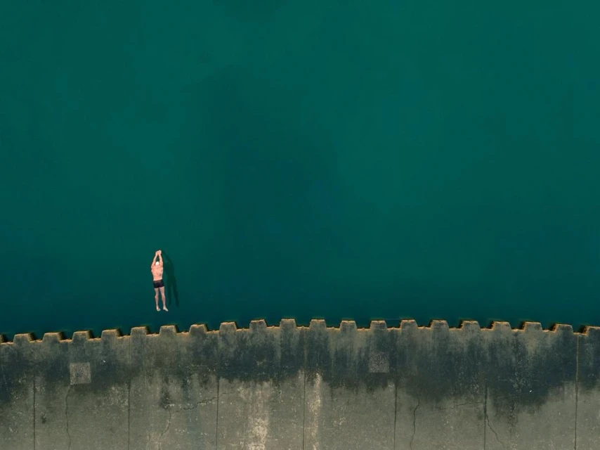 Aerial view of an adult male floating in turquoise water next to a concrete pier on a sunny day, showing tranquility and open water.