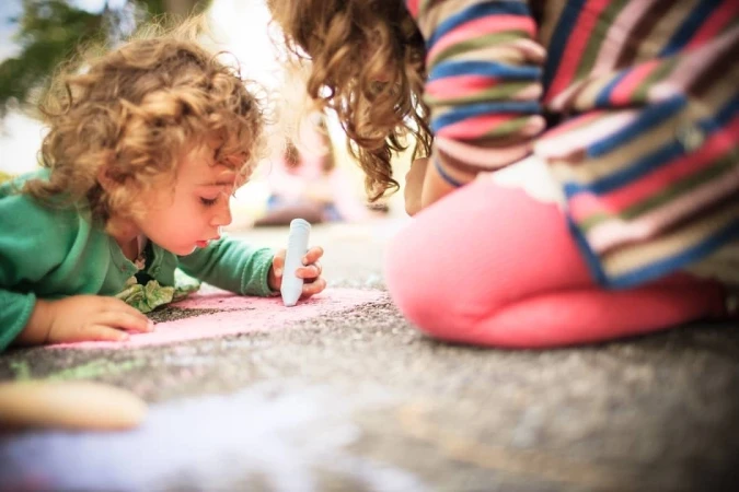 Child draws with chalk on pavement, outside, close-up with colorful chalk drawings and another girl in the background, sunny daytime.