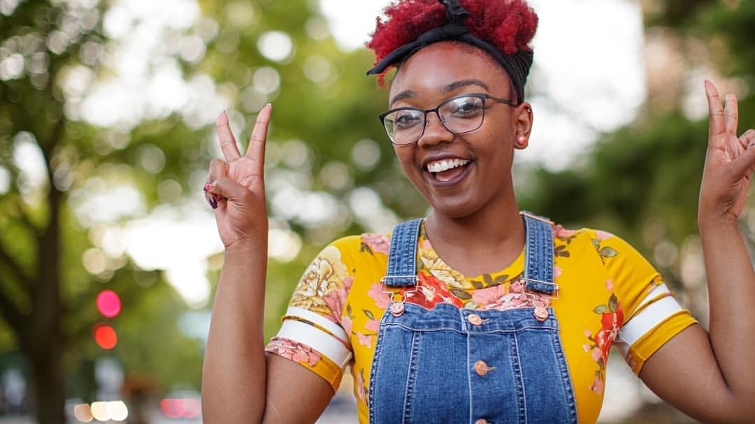 Smiling woman with glasses and burgundy hair outdoors makes peace sign with both hands, wearing a yellow floral shirt and overalls.