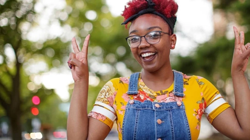 Smiling woman with glasses and burgundy hair outdoors makes peace sign with both hands, wearing a yellow floral shirt and overalls.