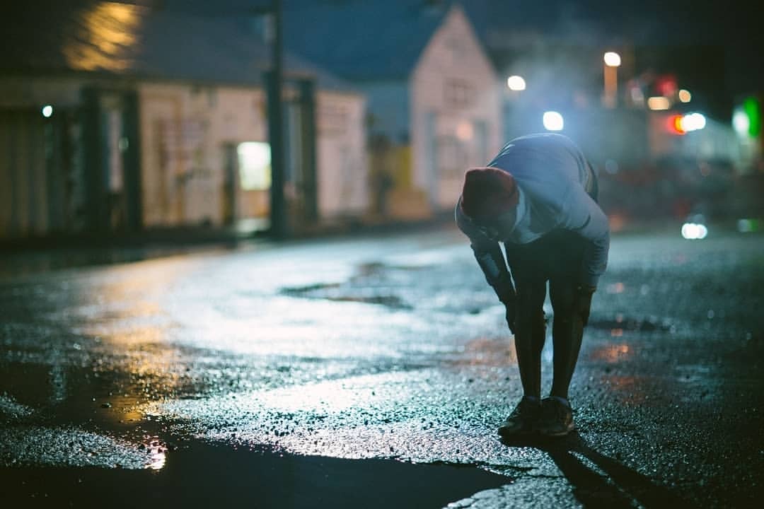 Adult male in athletic wear pauses on a wet road at night, leaning over with hands on knees, with buildings in the blurred background.