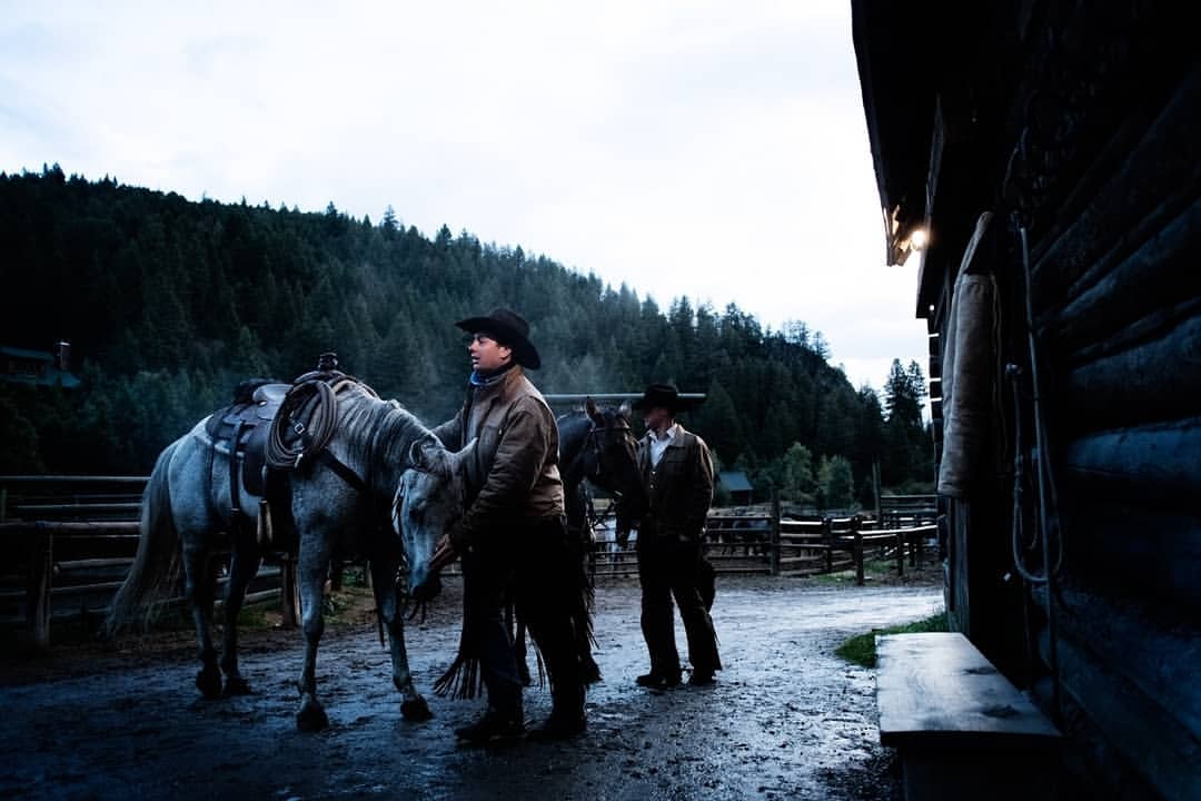 Two cowboys and their horses stand outside a log cabin on a ranch with a lush green forest and overcast sky in the background.