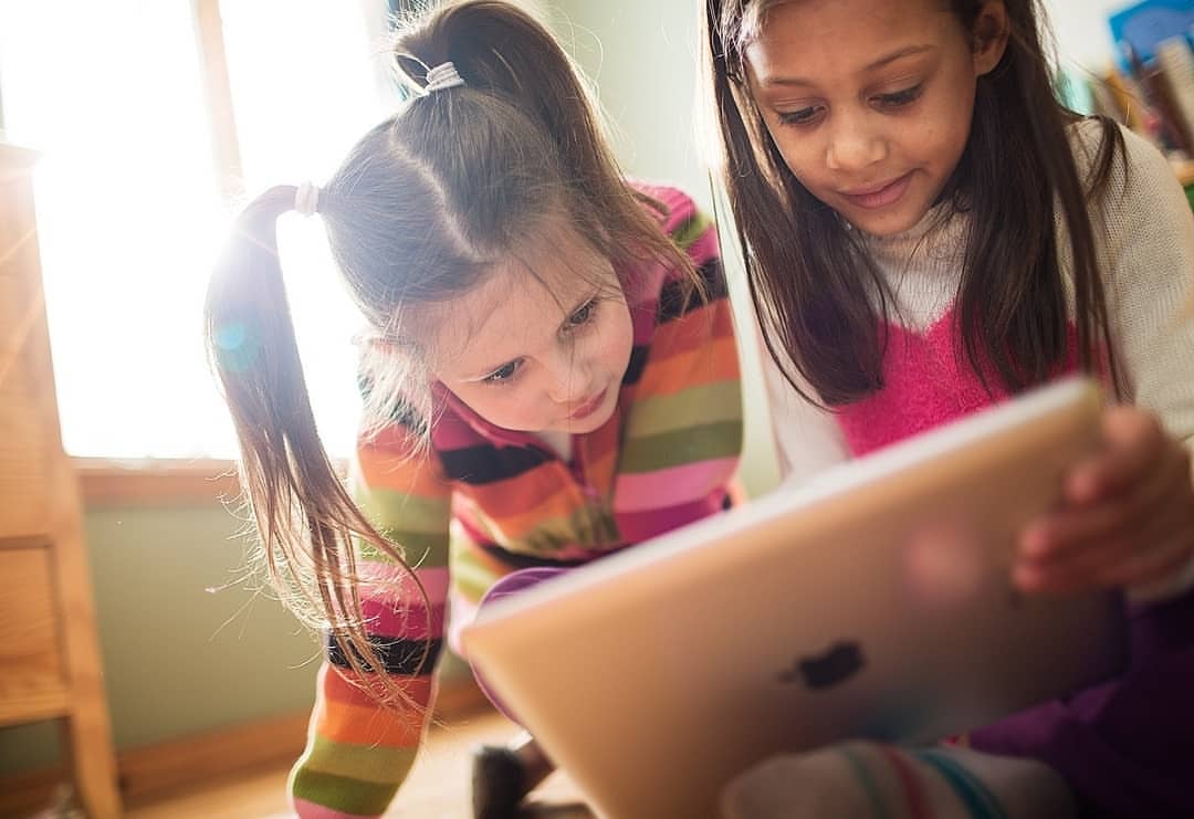 Two young girls share a tablet device indoors, bathed in bright light. One girl has pigtails and a striped shirt, the other has long hair.