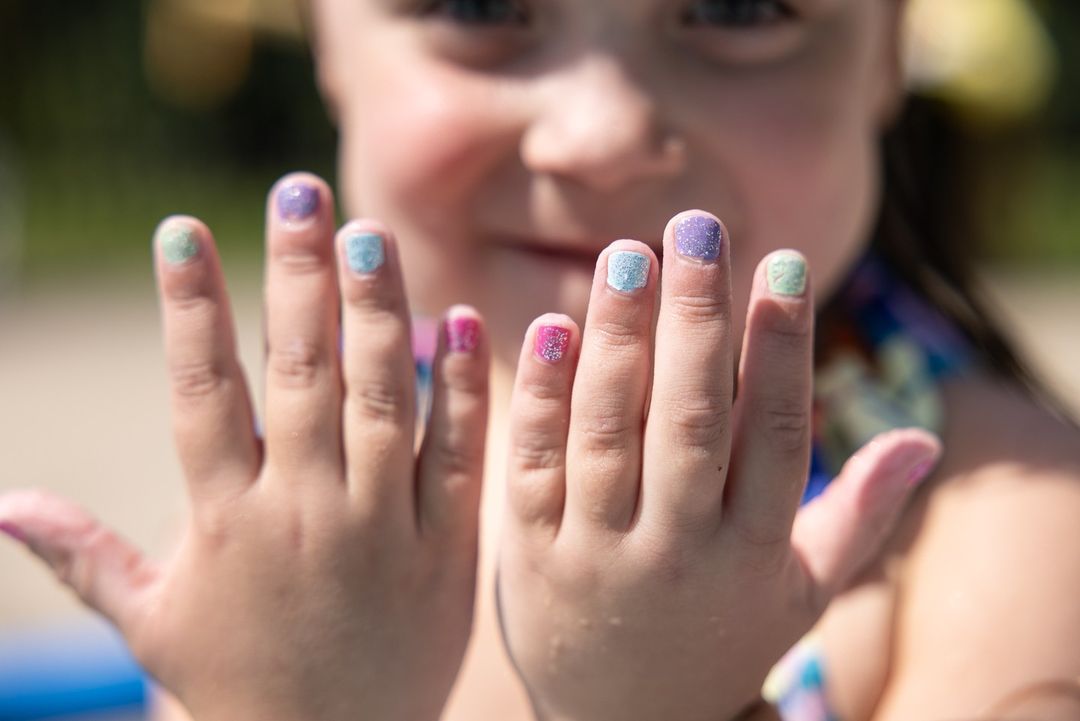 Close-up of a smiling child showing their hands, each fingernail painted a different pastel color with glitter, captured in an outdoor setting.