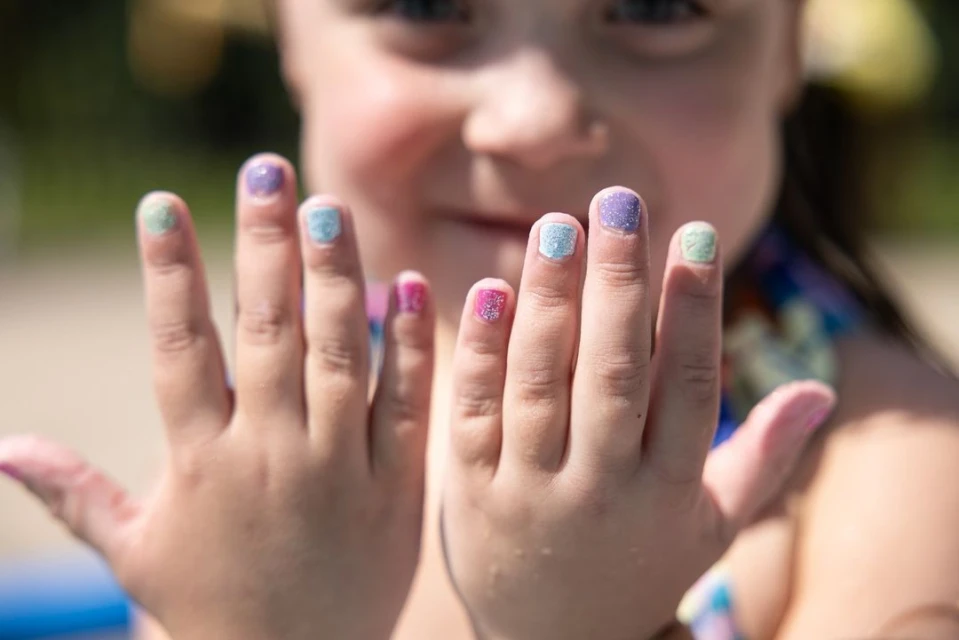 Close-up of a smiling child showing their hands, each fingernail painted a different pastel color with glitter, captured in an outdoor setting.