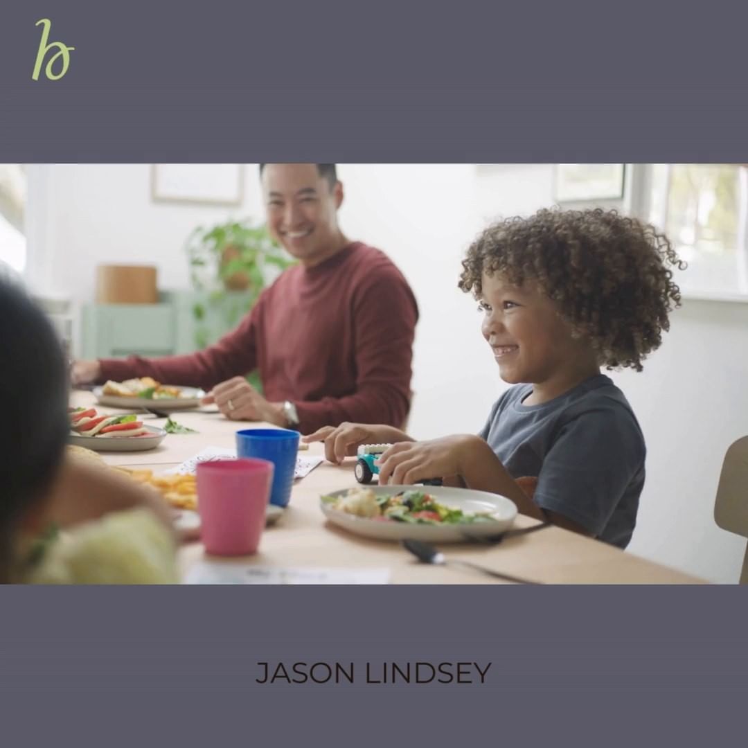 A family gathers around a table for a meal, featuring a smiling child and father, with dishes and drinks in a brightly lit interior.