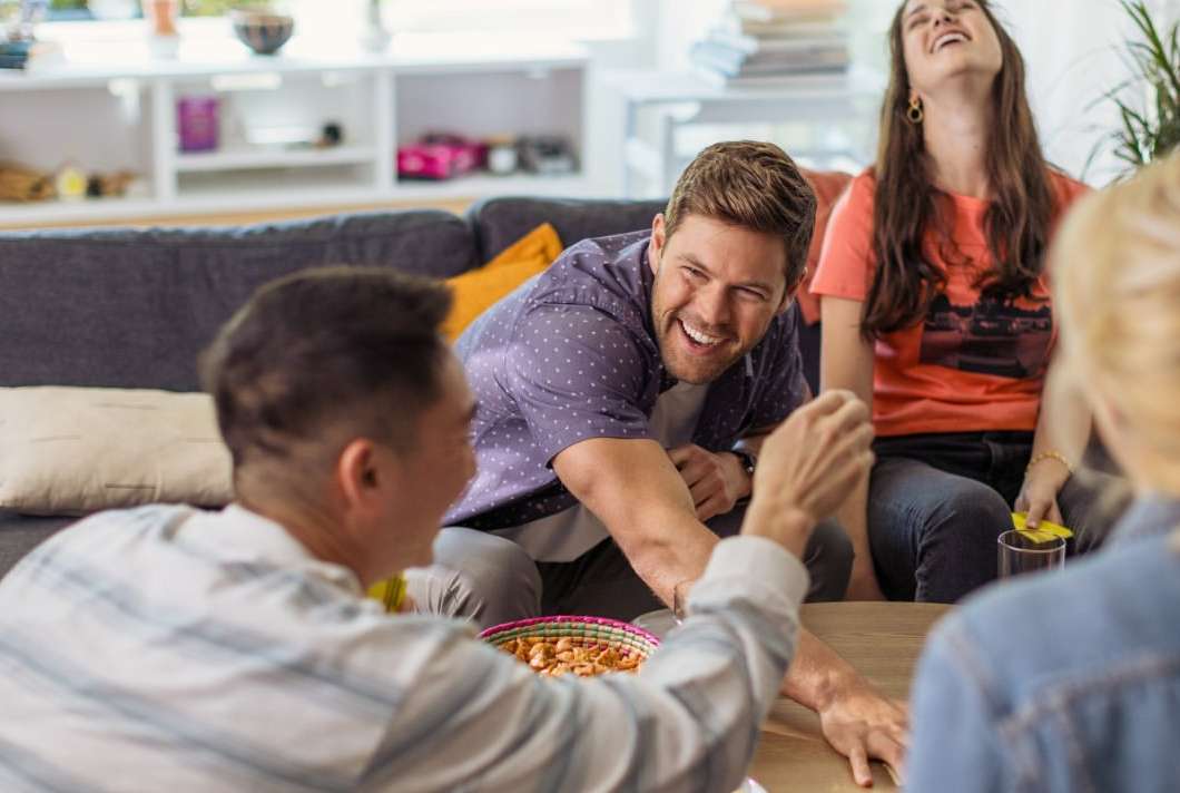 A group of friends laughing and socializing indoors. People gather around a table with snacks, enjoying each other's company in a casual setting.