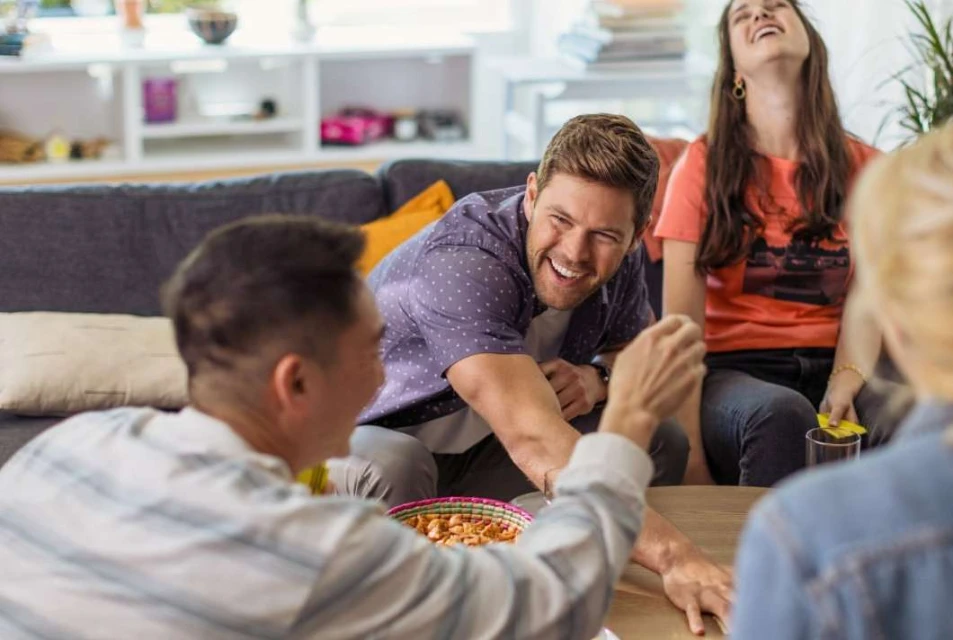 A group of friends laughing and socializing indoors. People gather around a table with snacks, enjoying each other's company in a casual setting.