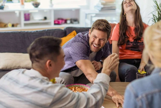 A group of friends laughing and socializing indoors. People gather around a table with snacks, enjoying each other's company in a casual setting.