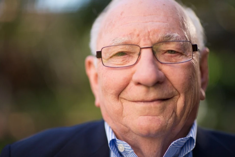 Close-up portrait of an elderly man with glasses, wearing a blue blazer and light blue collared shirt, smiling outdoors.