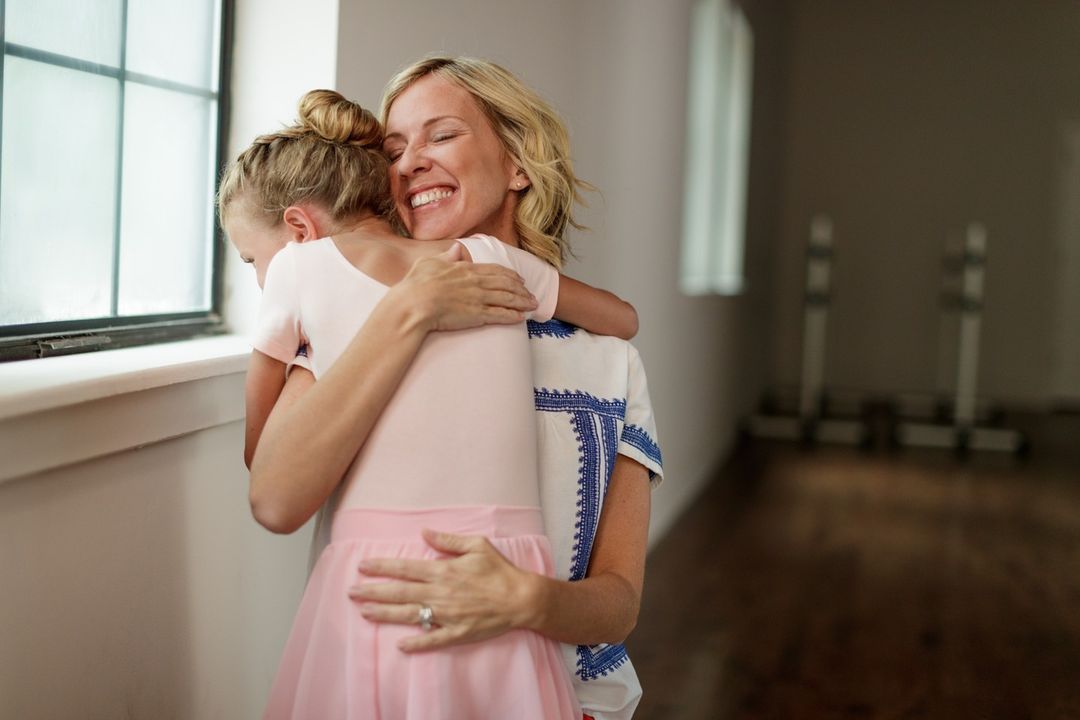 Indoor shot of a blonde adult female hugging a child in a pink ballet outfit next to a window in a bright room, showing a happy, affectionate moment.