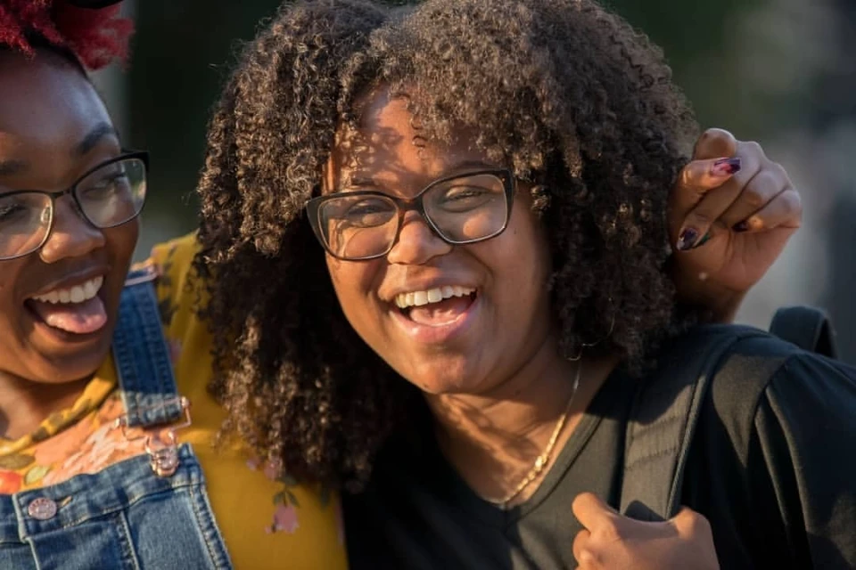 Two smiling young women with curly hair, one in denim overalls and a yellow shirt, and the other in a black t-shirt outdoors.