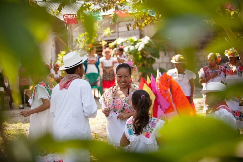 A group of people in traditional clothing gather outdoors, smiling and interacting, with festive decorations visible in the background.