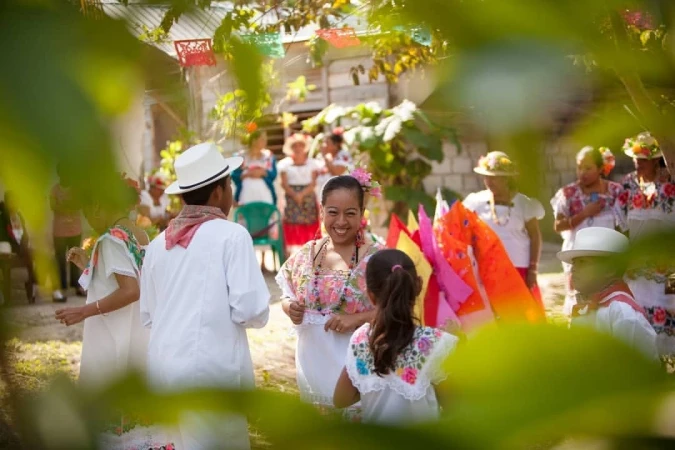 A group of people in traditional clothing gather outdoors, smiling and interacting, with festive decorations visible in the background.