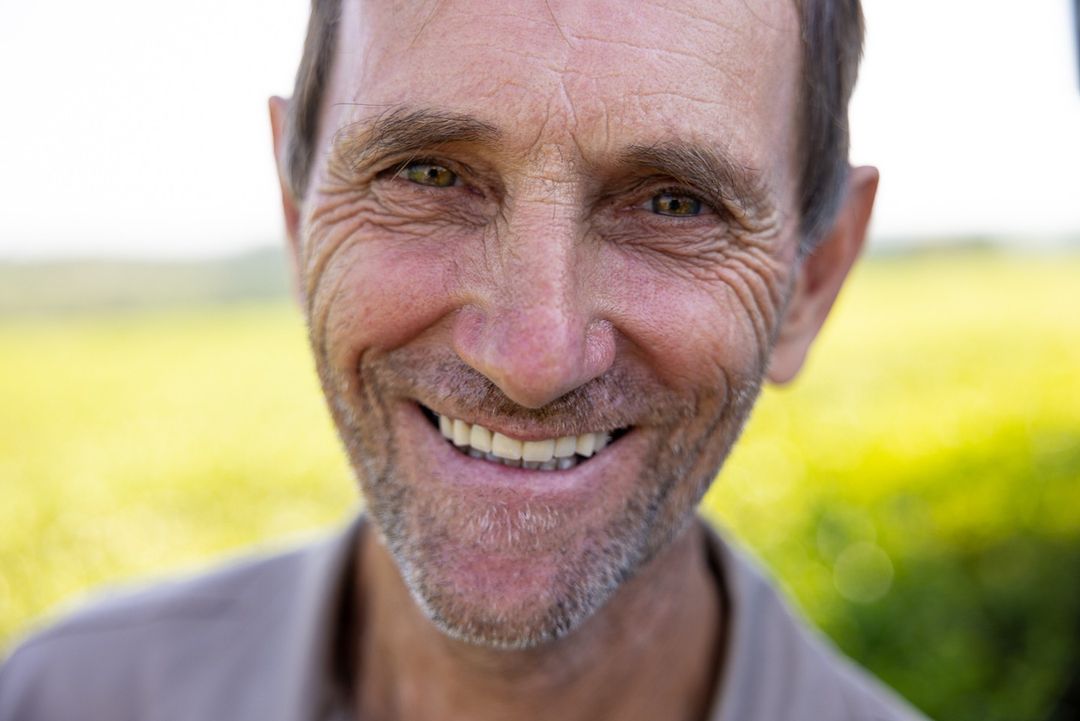 Close-up of an elderly adult male with green eyes and a big smile, captured in soft, natural light, against an out-of-focus green background.