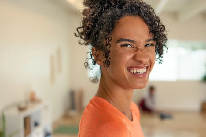 Close-up portrait of a smiling young woman with curly hair wearing an orange top in a bright interior space.