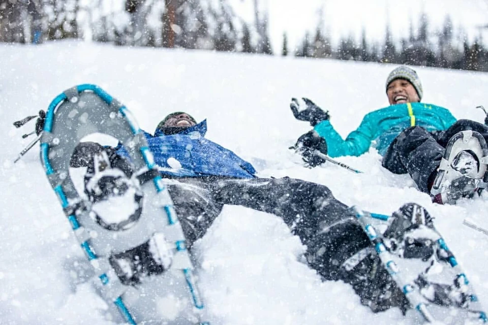 Two people in winter clothing enjoy a snowy day, laying in the snow with snowshoes in the foreground and trees in the background.