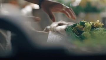 Close-up shot of a hand arranging a vibrant bouquet with yellow flowers wrapped in green paper. Soft, diffused light emphasizes the textures.
