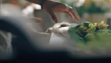 Close-up shot of a hand arranging a vibrant bouquet with yellow flowers wrapped in green paper. Soft, diffused light emphasizes the textures.