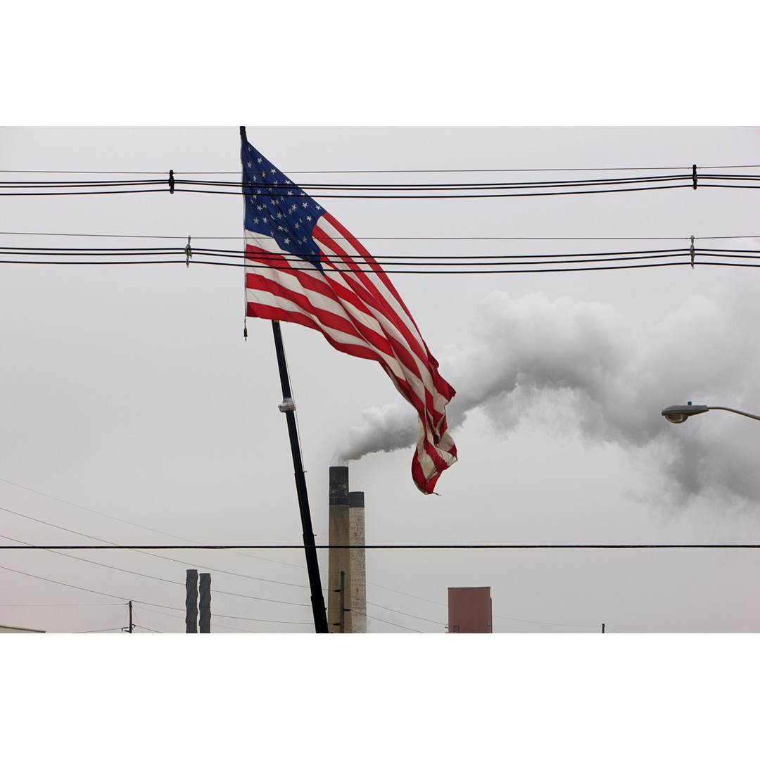 American flag waving in an outdoor scene with industrial elements, including smoke stacks and power lines, against an overcast sky.