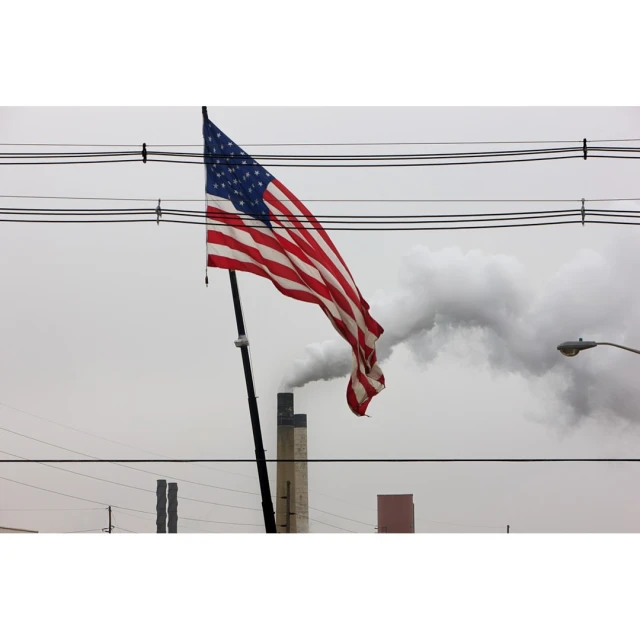 American flag waving in an outdoor scene with industrial elements, including smoke stacks and power lines, against an overcast sky.