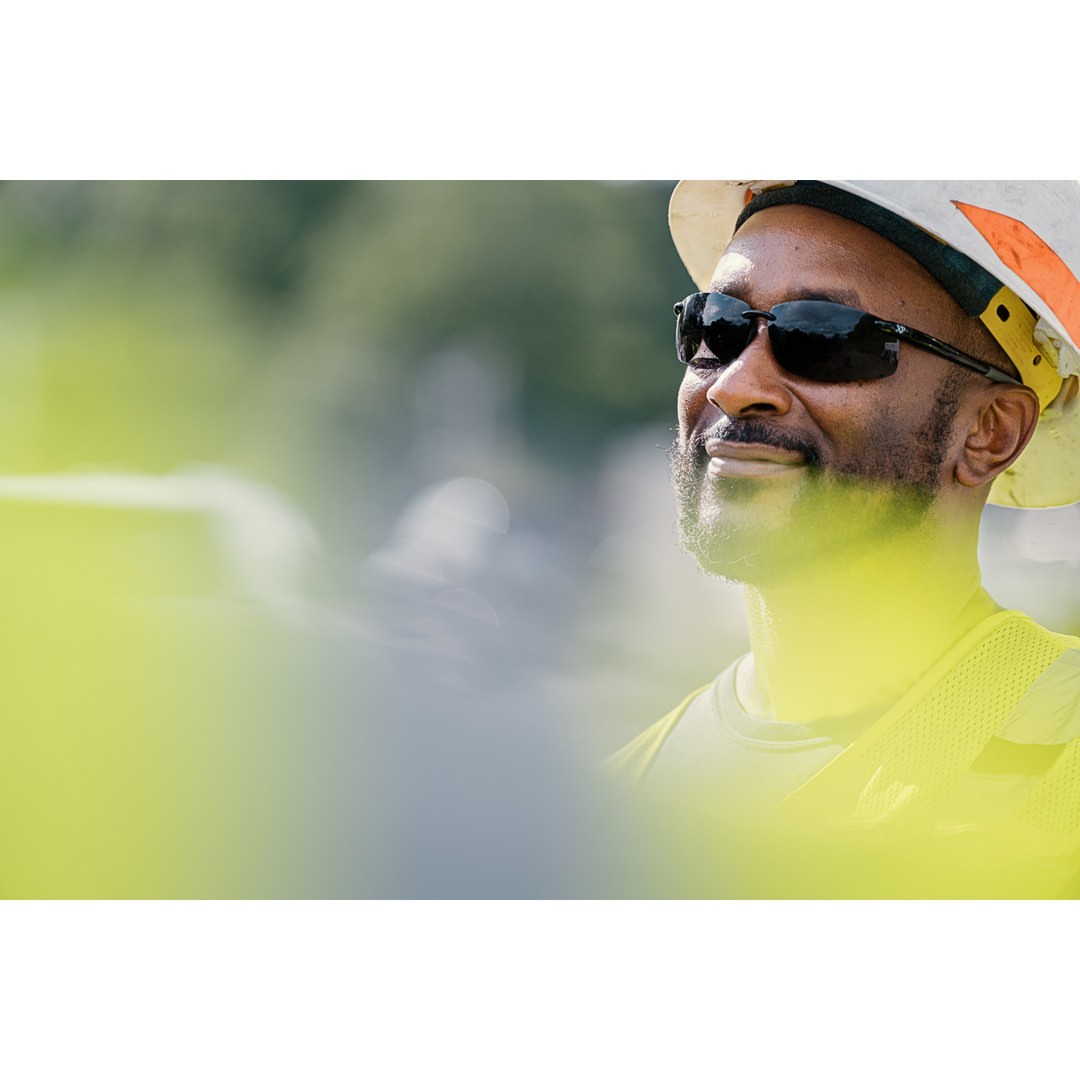 Close-up of a smiling construction worker with a white hard hat, safety glasses, and a high-visibility vest in an outdoor setting.